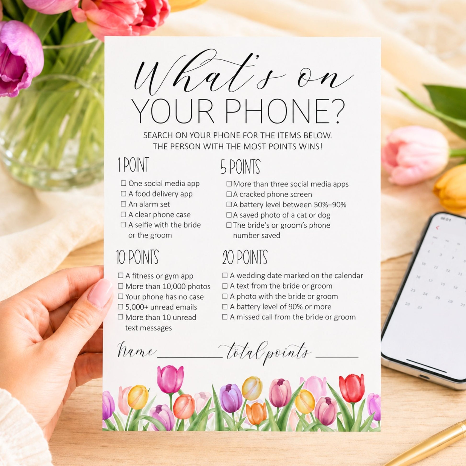 A bridal shower game card titled 'What's on your phone?' held by a hand over a floral table setting with a notebook nearby.