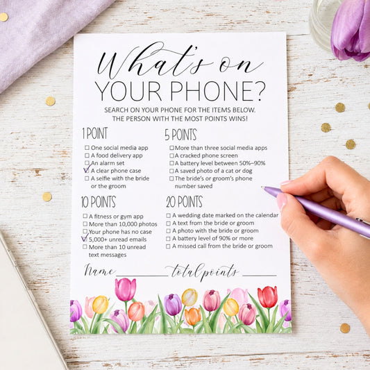 A printed bingo game card titled "What's on your phone?" with checklist questions and a tulip floral border, photographed on a white wood surface with a hand holding a purple pen over it. A pink fabric and a glass of purple drink are partially visible around the scene.