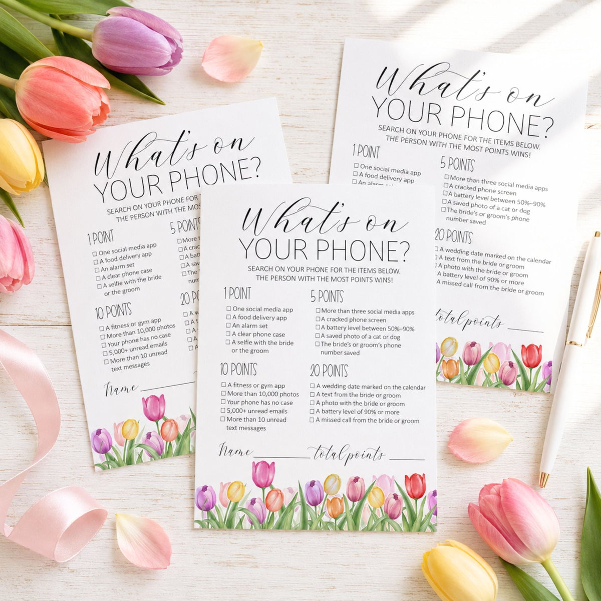 Three printed wedding favor cards titled 'What's on YOUR PHONE?' arranged on a white wood surface with tulips around the edges. The cards feature black script and serif text with a floral tulip border at the bottom and a scoring system with points and gift suggestions.
