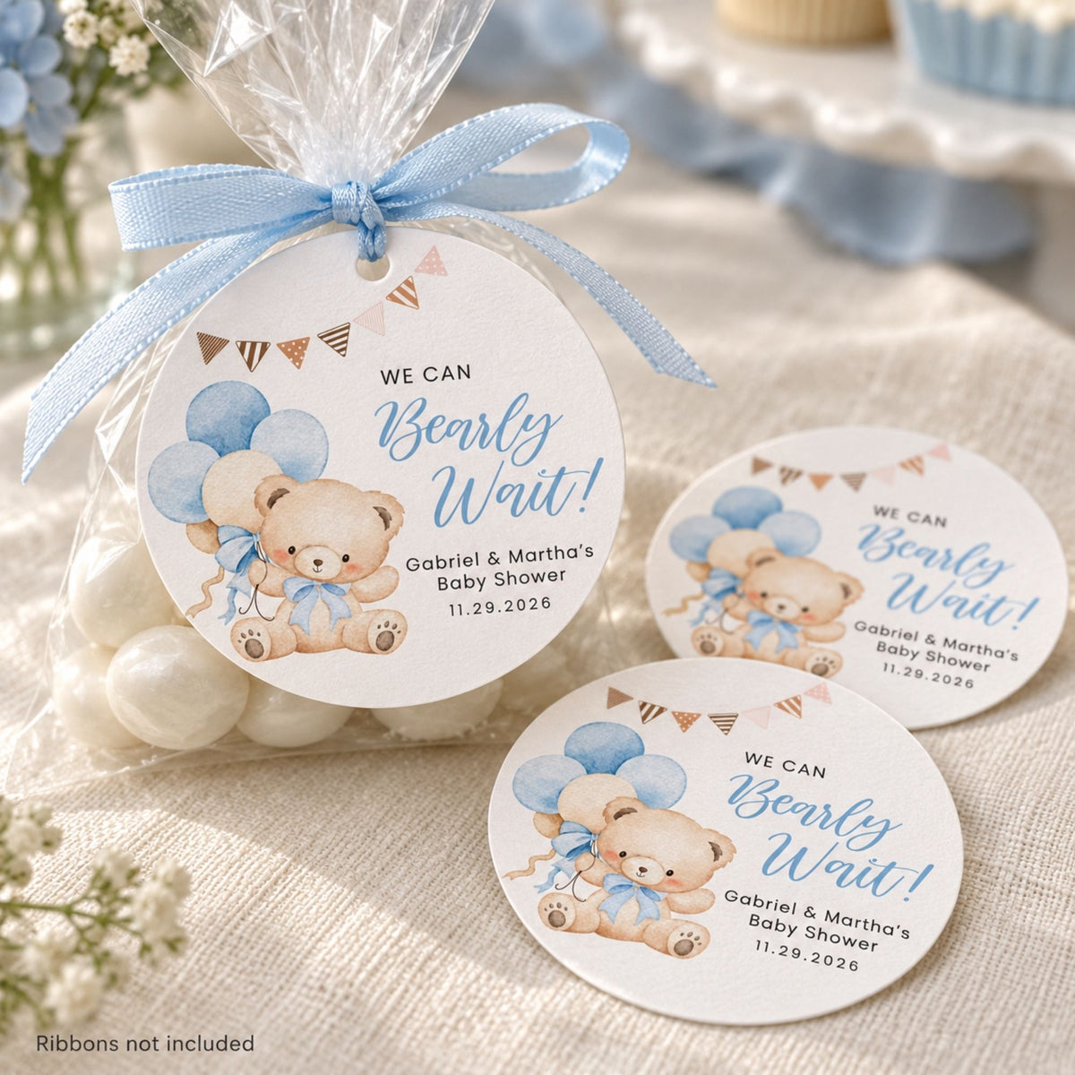Three round baby shower favor tags with teddy bear illustrations and blue ribbon bows are shown on a clear candy bag and on a fabric surface. The tags read 'WE CAN Bearly Wait!' with personalized names and date.