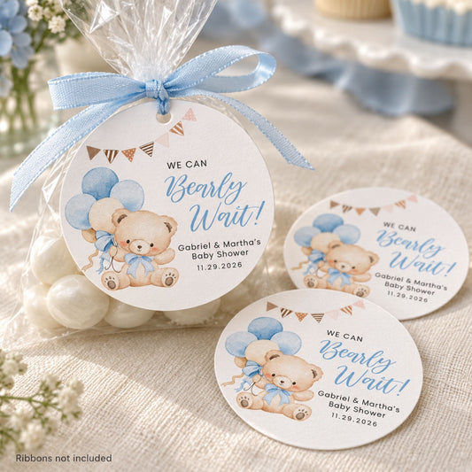 Three round baby shower favor tags with teddy bear illustrations and blue ribbon bows are shown on a clear candy bag and on a fabric surface. The tags read 'WE CAN Bearly Wait!' with personalized names and date.
