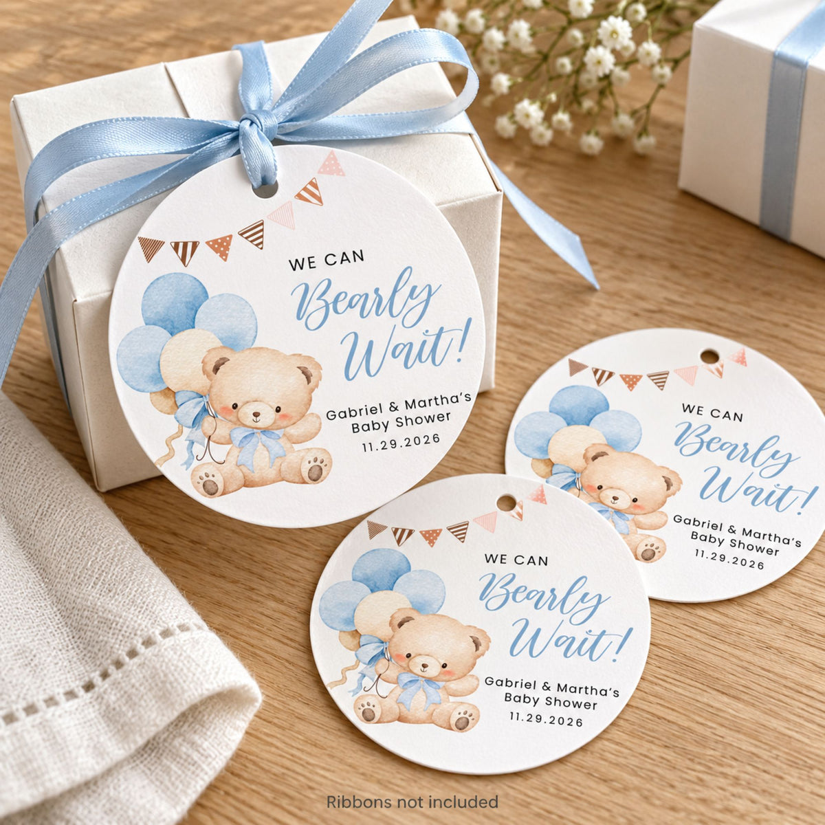 Personalized baby shower favor tags with teddy bear illustrations and blue balloons, shown attached to a gift box and placed on a wooden surface with a ribbon.