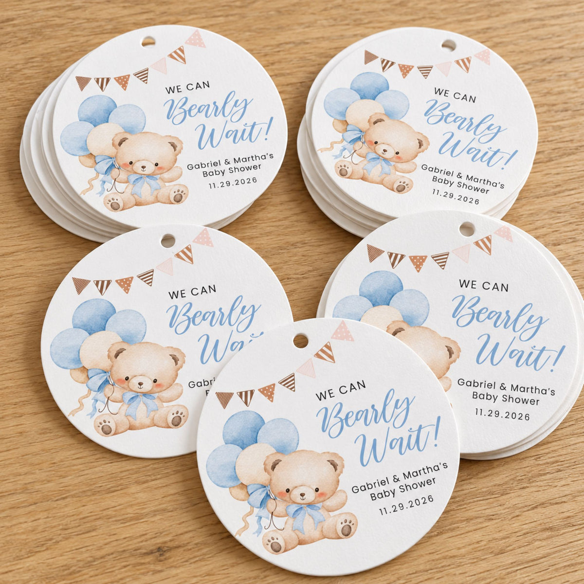 A set of round white baby shower favor tags featuring a watercolor teddy bear, blue balloons, bunting flags, and the text 'WE CAN Bearly Wait!' with personalized names and date printed below, arranged on a light wood surface.