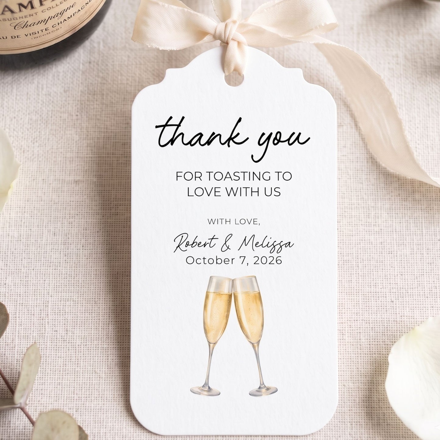 A personalized wedding thank-you gift tag with a cream ribbon bow, featuring the text 'thank you FOR TOASTING TO LOVE WITH US' and an illustration of two champagne glasses, styled on a linen surface with a champagne bottle partially visible.
