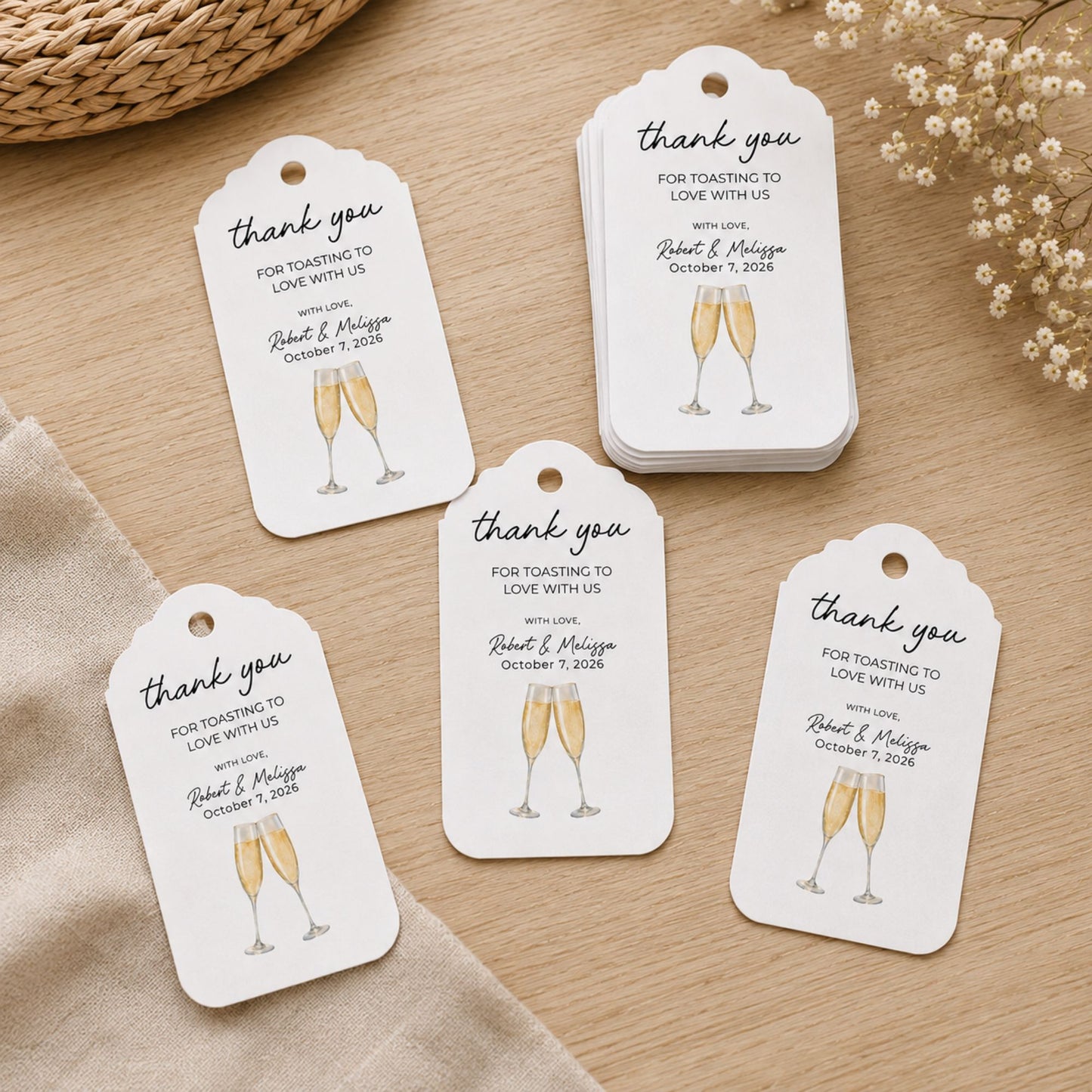A stack of personalized wedding toast tags with matching individual tags displayed on a light wood surface, styled with a woven basket, beige fabric, and dried flowers.