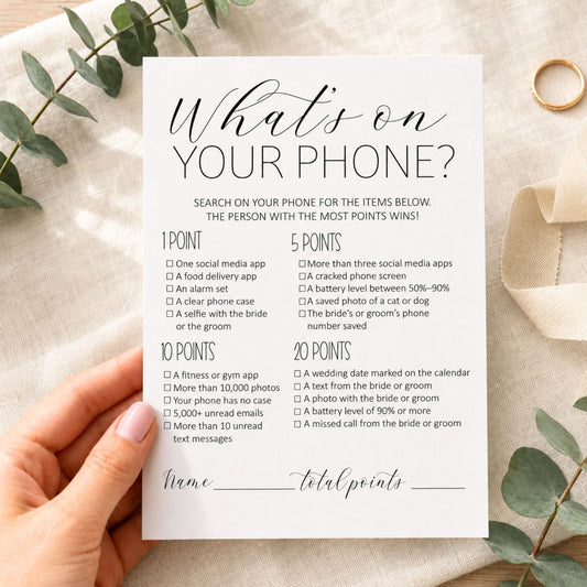 A hand holds a white printed card titled "What's on YOUR phone?" with lists of phone-related items and scoring points, styled on a light fabric background with eucalyptus leaves and a gold ring.