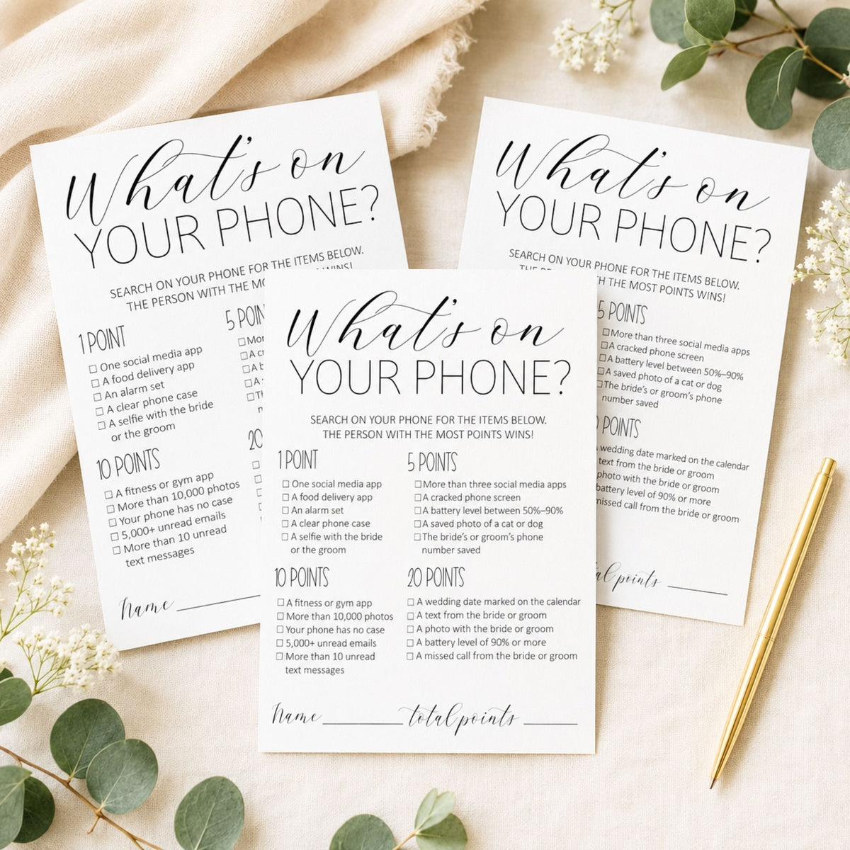 Three white printed game cards titled "What's on YOUR phone?" arranged on a light neutral surface with scattered white flowers and green leaves. The cards feature black script and block text with numbered clues and a total points field, plus a gold pen placed at the bottom right.