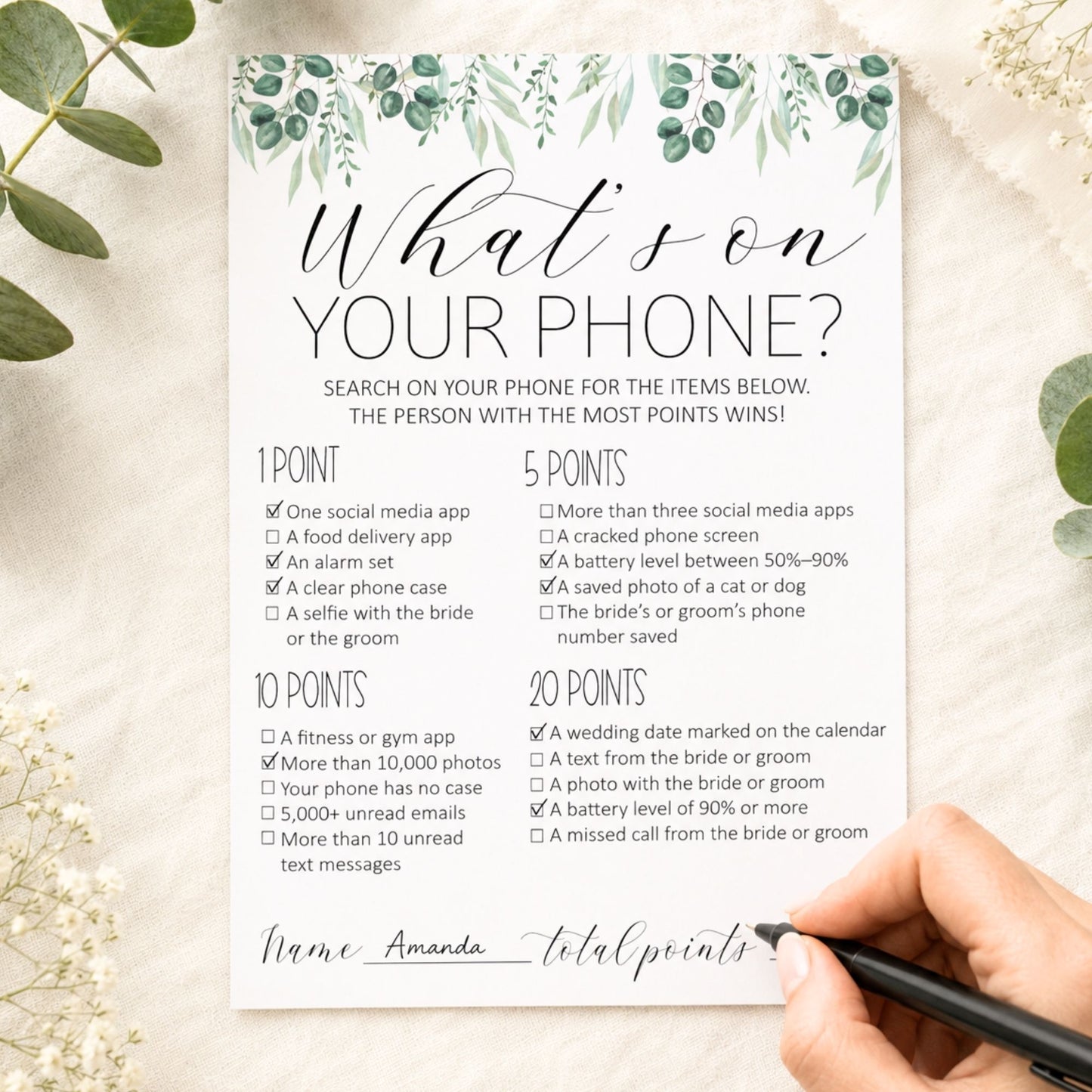 A printed party game card titled "What's on YOUR PHONE?" with illustrated greenery at the top, shown on a light fabric background with a hand holding a black marker. The card lists checked boxes for phone apps, photos, emails, and other phone-related items, with a blank line for total points.