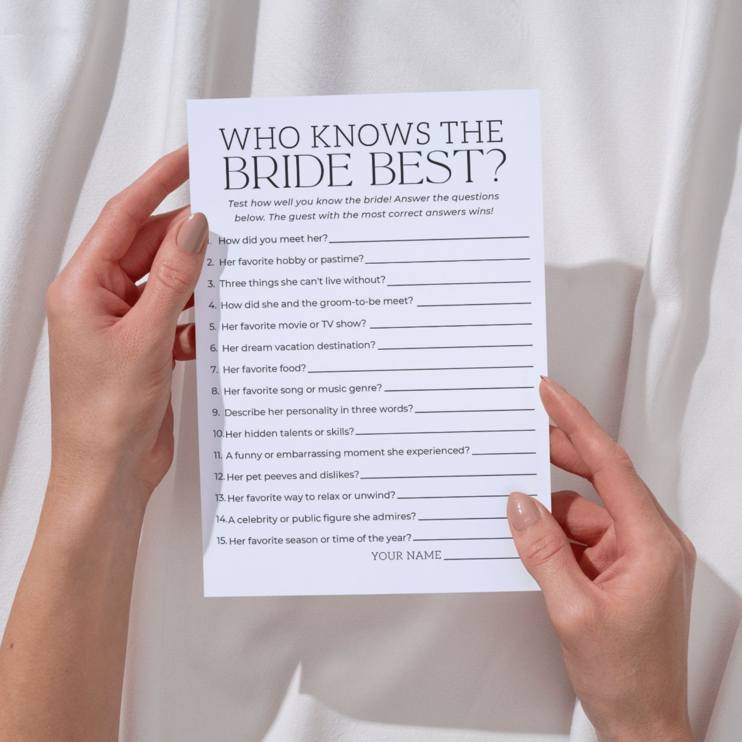 A person holding a white bridal shower game card titled "WHO KNOWS THE BRIDE BEST?" against a soft white fabric background. The card features 15 numbered questions with blank lines for answers, including "How did you meet her?", "Her favorite food?", and "Her dream vacation destination?"
