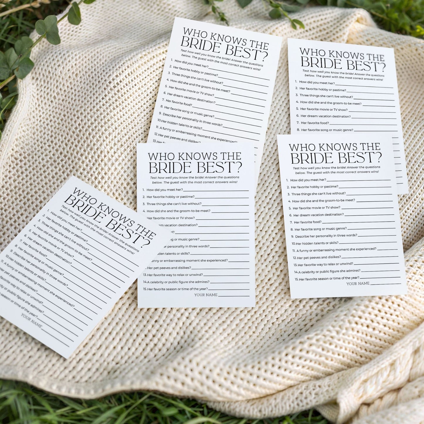 Multiple "Who Knows the Bride Best?" game cards scattered artistically on a cream-colored textured knit blanket outdoors on green grass. The image highlights the physical cards in a natural, celebratory setting.