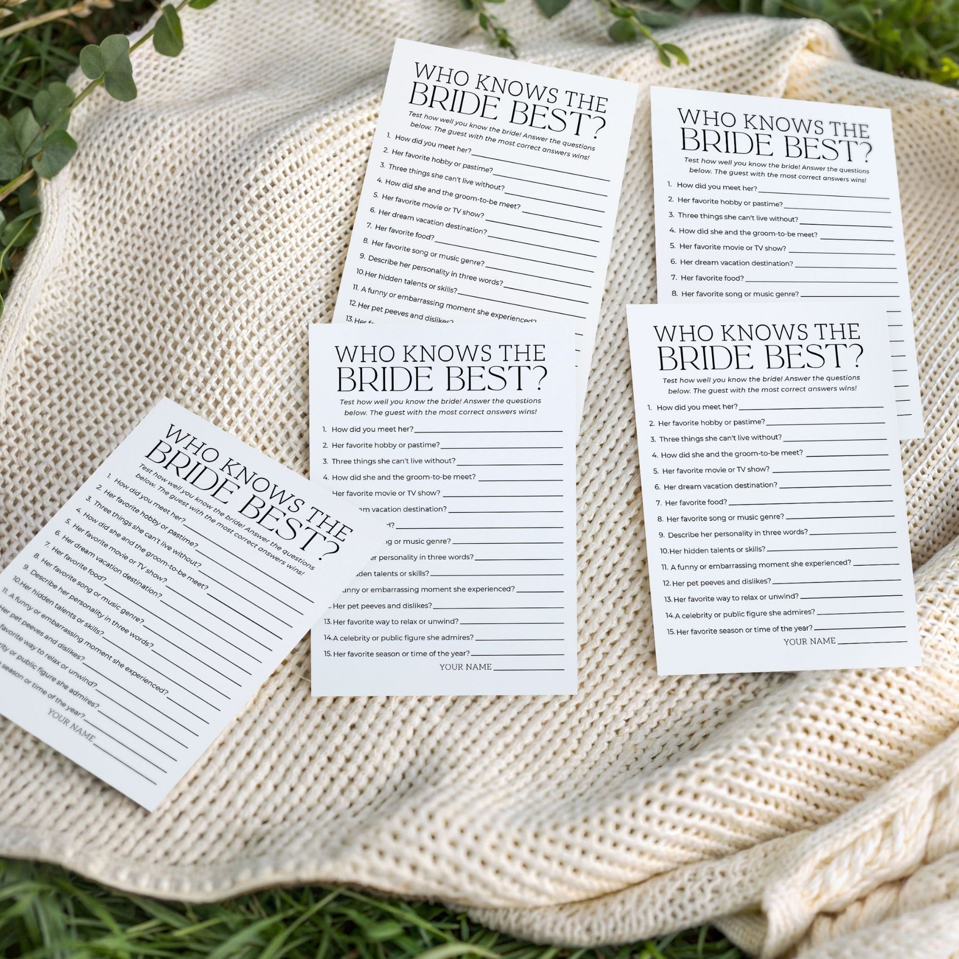 Multiple "Who Knows the Bride Best?" game cards scattered artistically on a cream-colored textured knit blanket outdoors on green grass. The image highlights the physical cards in a natural, celebratory setting.