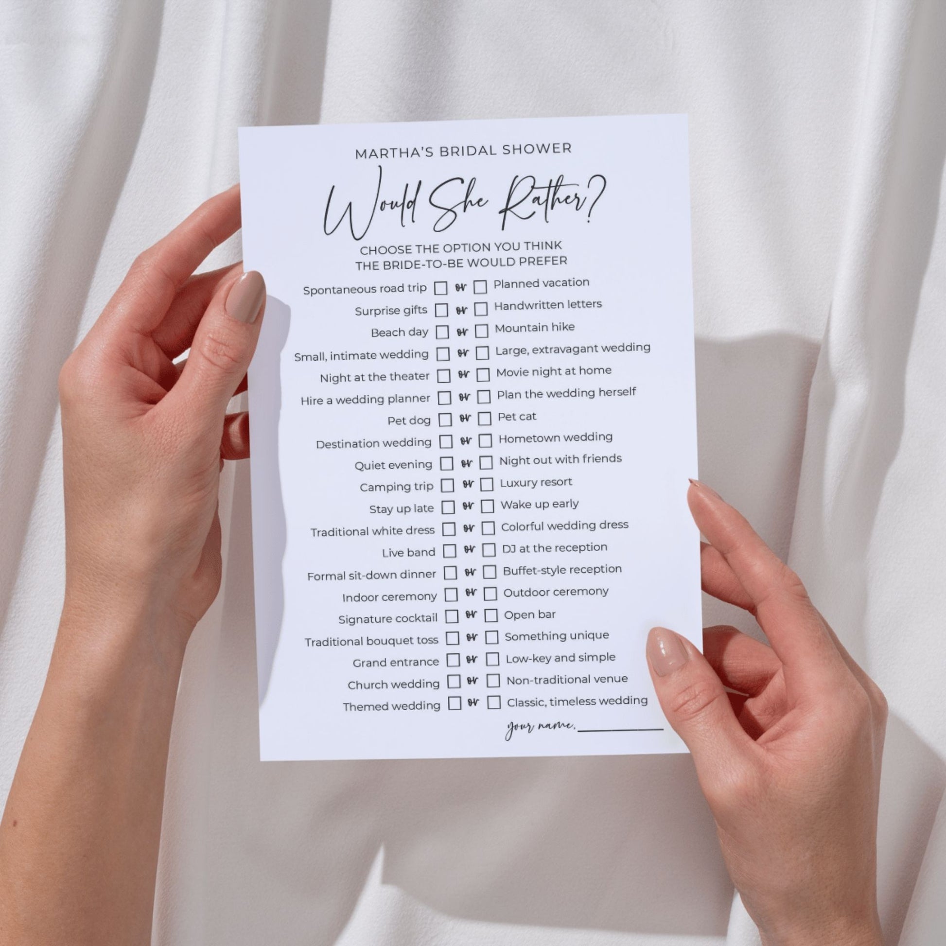 Hands with neutral manicured nails hold a 5x7 bridal shower game card against a draped white fabric background. The card is titled "Martha's Bridal Shower: Would She Rather?" in elegant black script. It features 20 "this or that" options with checkboxes, such as "Spontaneous road trip OR Planned vacation" and "Beach day OR Mountain hike," with a "your name" signature line at the bottom.