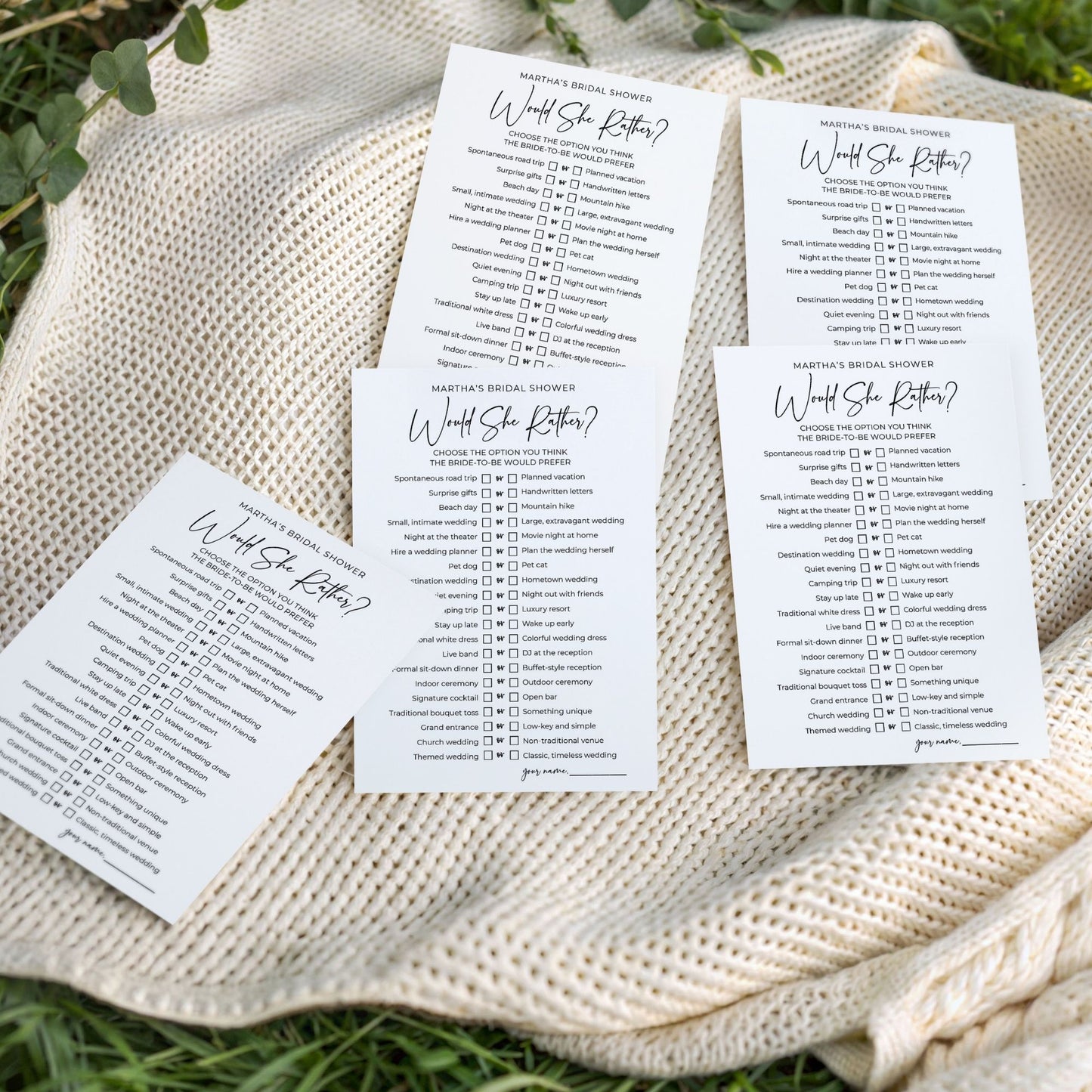 A flat-lay photo of five "Would She Rather" bridal shower game cards fanned out on a cozy, cream-colored cable-knit blanket. The blanket is spread over green grass in bright, natural outdoor light. The minimalist white cards feature clean black text, displaying a "this or that" list of fun wedding-themed questions.