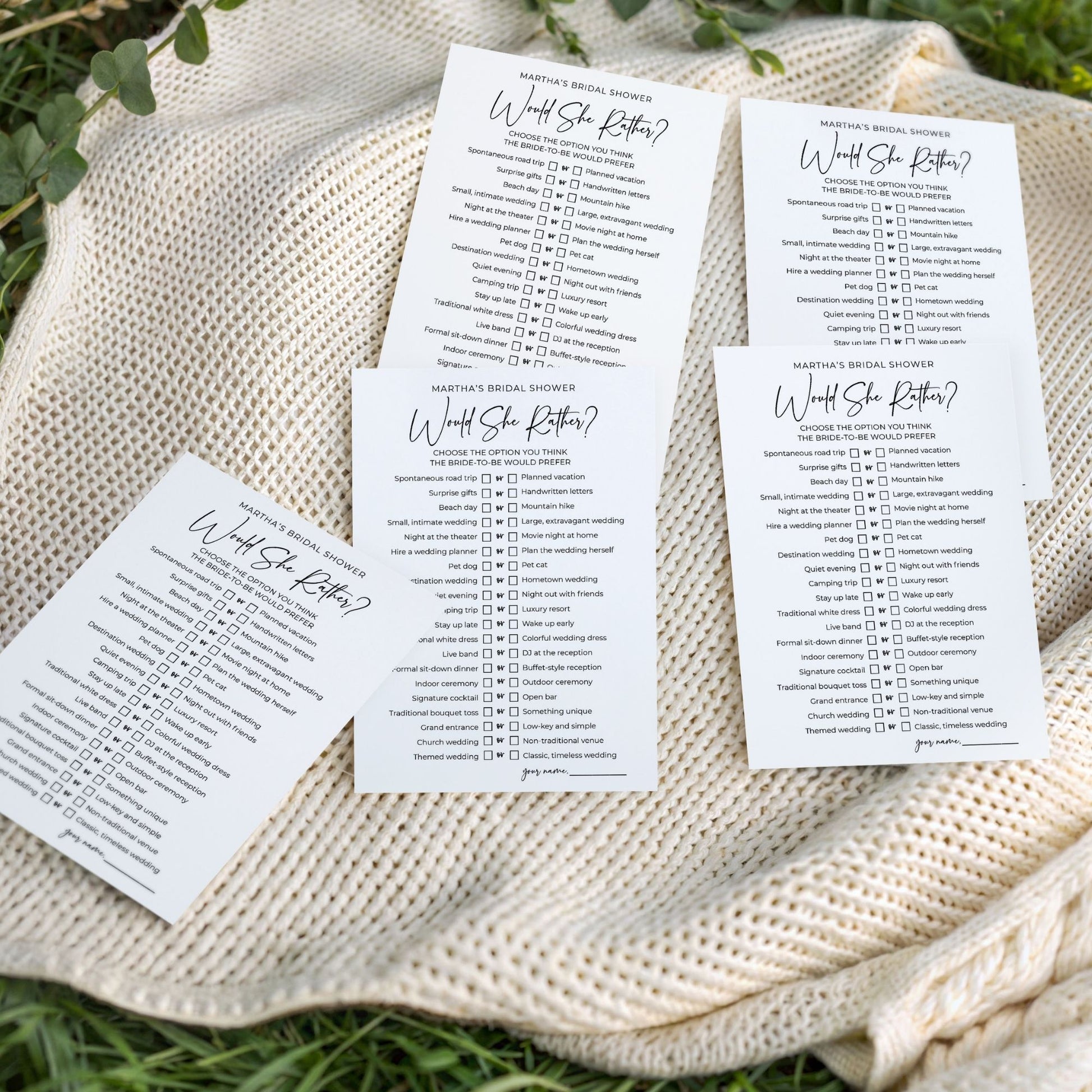 A flat-lay photo of five "Would She Rather" bridal shower game cards fanned out on a cozy, cream-colored cable-knit blanket. The blanket is spread over green grass in bright, natural outdoor light. The minimalist white cards feature clean black text, displaying a "this or that" list of fun wedding-themed questions.