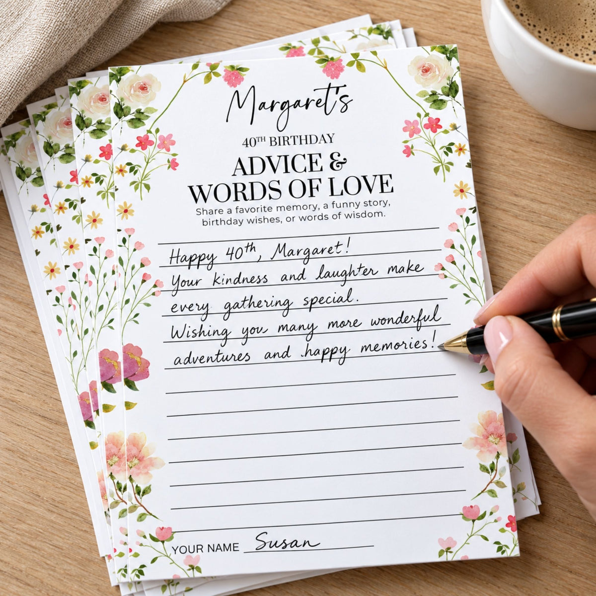 A stack of personalized floral birthday cards on a wooden table, with a hand holding a black pen writing on one card. A coffee cup is partially visible in the top right corner.