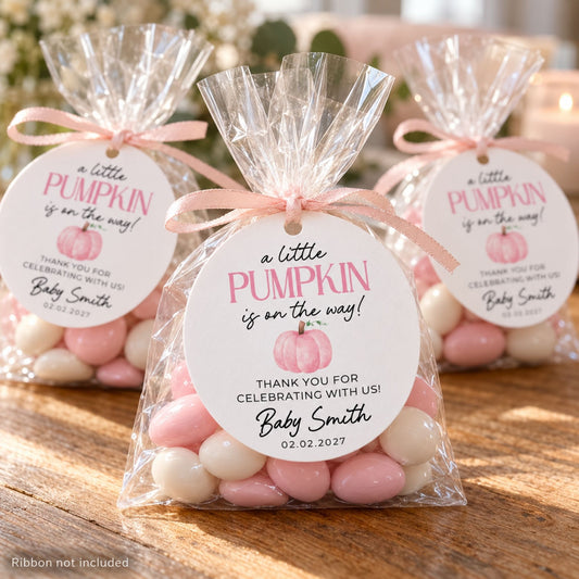 Three clear cellophane favor bags filled with pastel pink and cream candies, tied with pale pink ribbons, and each featuring a round printed tag that reads 'a little PUMPKIN is on the way!' and 'Baby Smith.' The bags are arranged on a wooden surface with a softly blurred outdoor background.