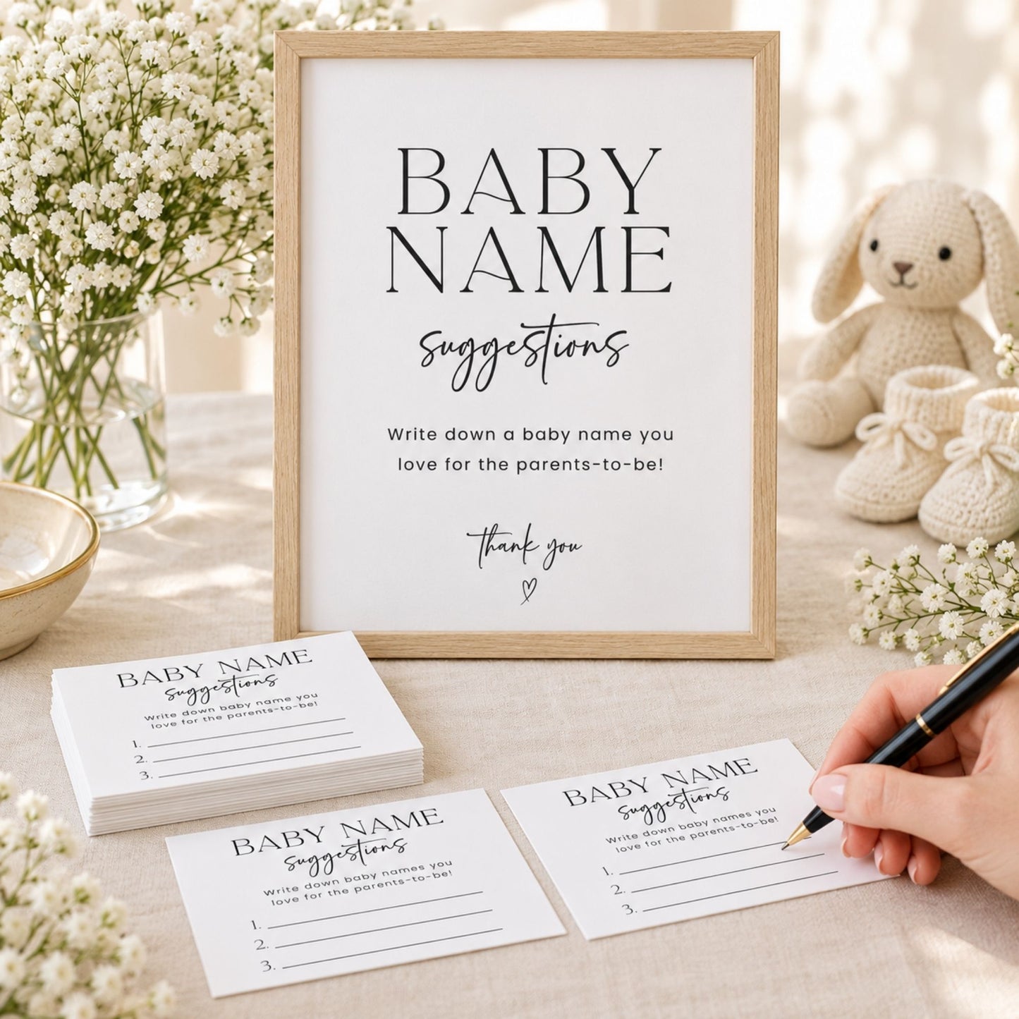 A framed baby name suggestion sign stands on a light fabric surface, with matching printed cards stacked in front and a hand holding a pen over one card. The scene is styled with baby flowers, a knitted bunny toy, and soft natural light.