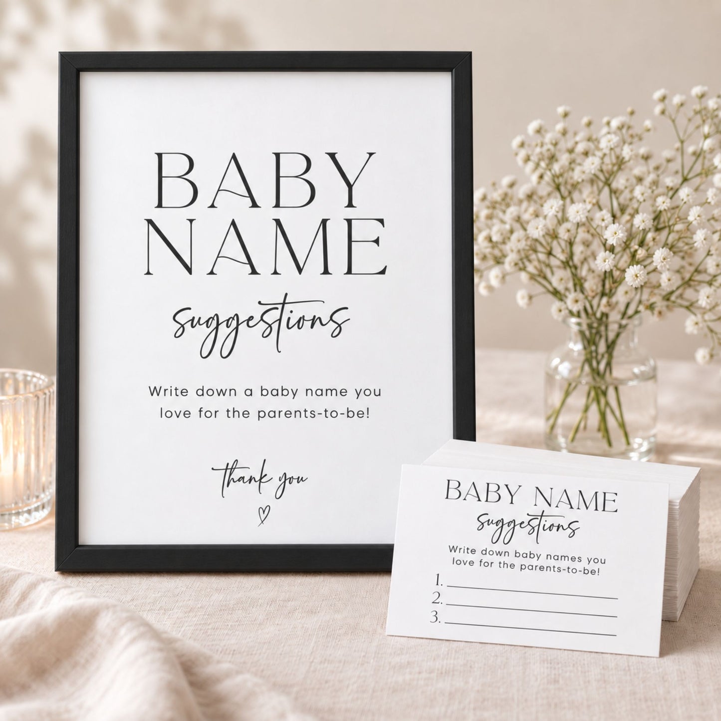 A black-framed baby name suggestions printout and matching printed cards are styled on a light fabric surface with a glass candle, baby's breath flowers, and a neutral background.