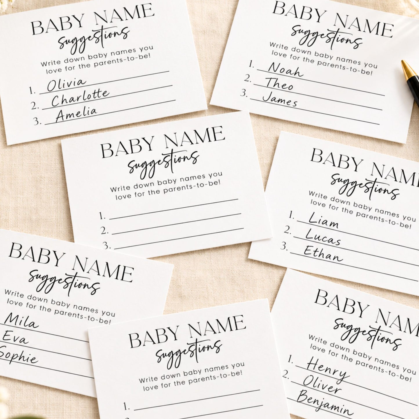 A set of printed baby name suggestions cards laid out on a light fabric background, with handwritten names visible on several cards and a small gold pen at the top right.