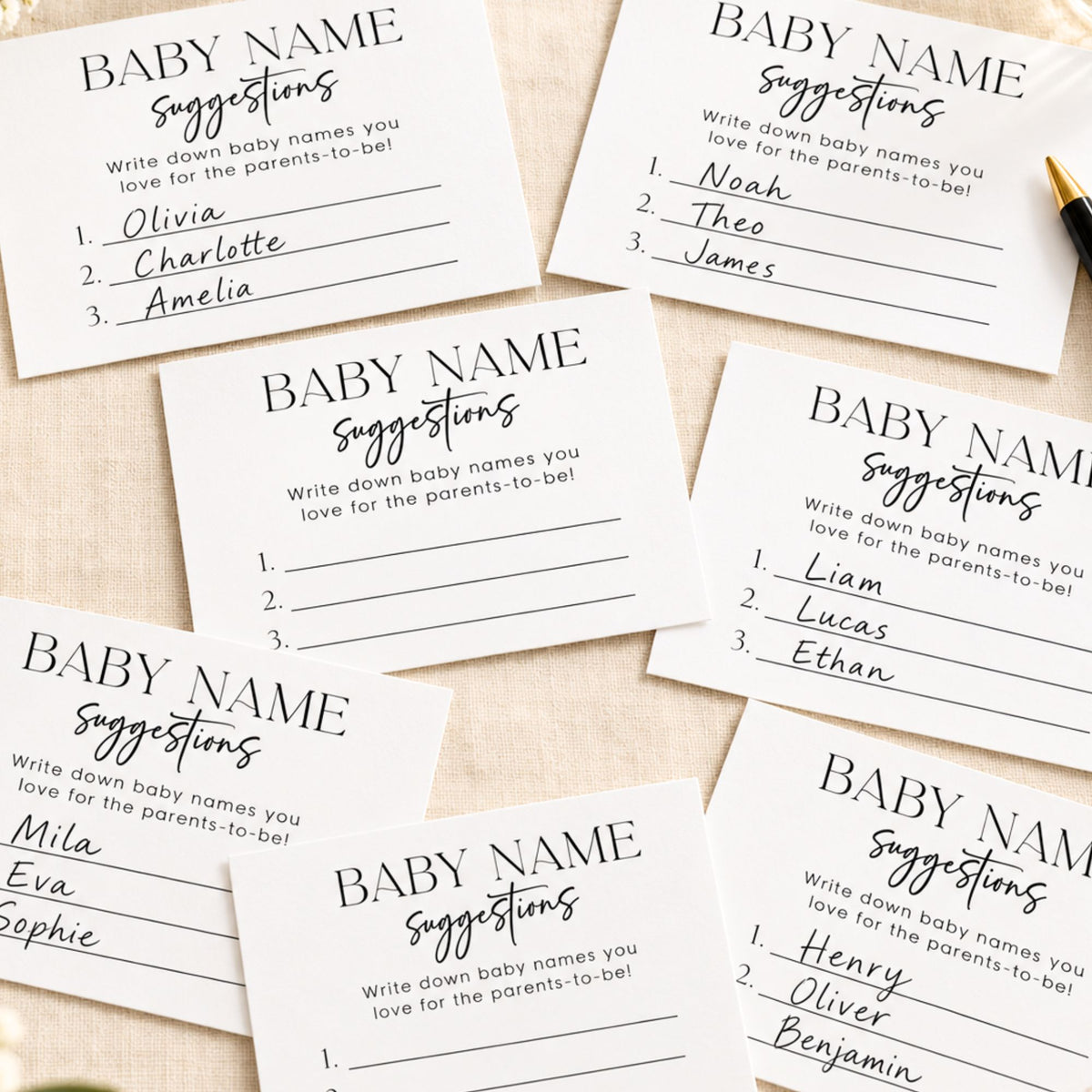 A set of printed baby name suggestions cards laid out on a light fabric background, with handwritten names visible on several cards and a small gold pen at the top right.