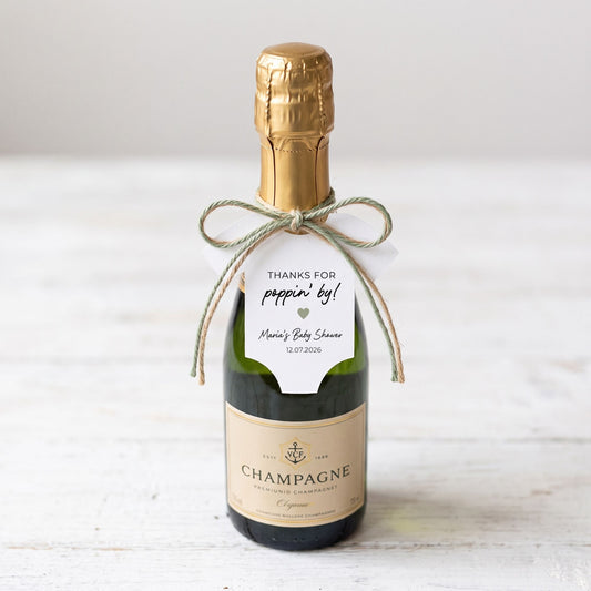 A personalized champagne bottle favor with a gold foil top, a beige label, and a twine bow holding a white thank-you tag, set on a white wooden surface.