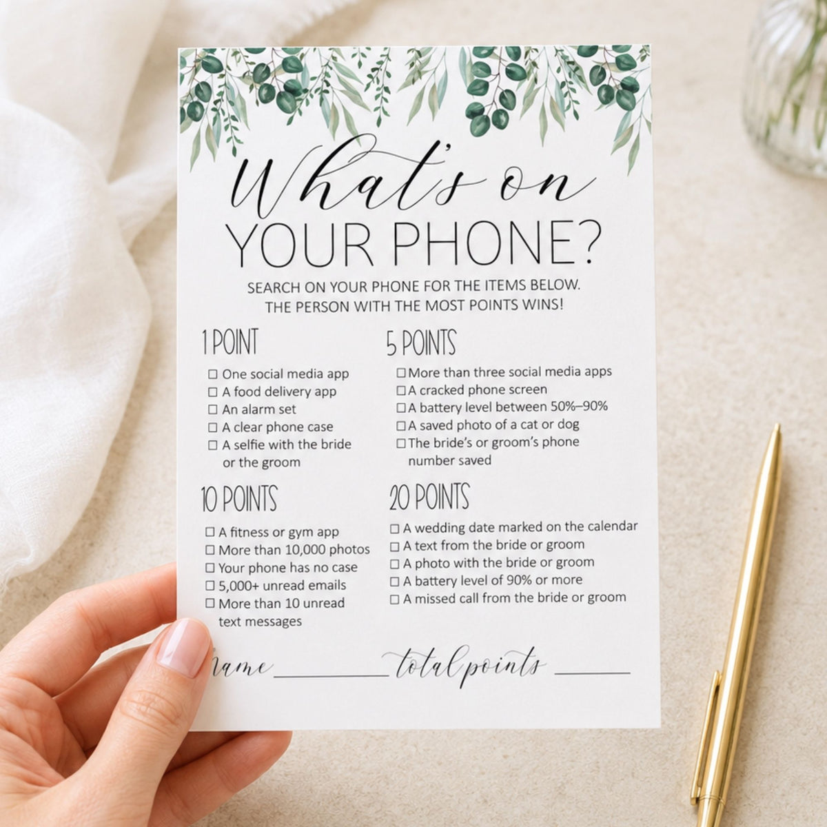 A hand holds a white printable card titled "What's on YOUR PHONE?" with green leafy illustrations at the top, surrounded by a gold pen and a glass bottle on a light fabric background.