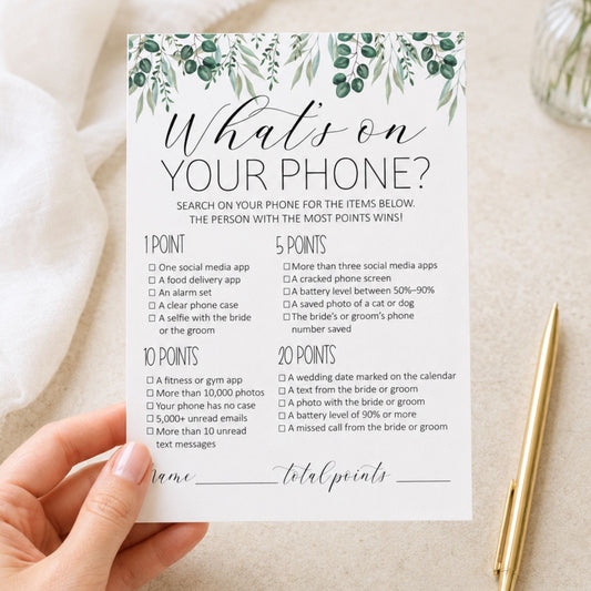 A hand holds a white printable card titled "What's on YOUR PHONE?" with green leafy illustrations at the top, surrounded by a gold pen and a glass bottle on a light fabric background.
