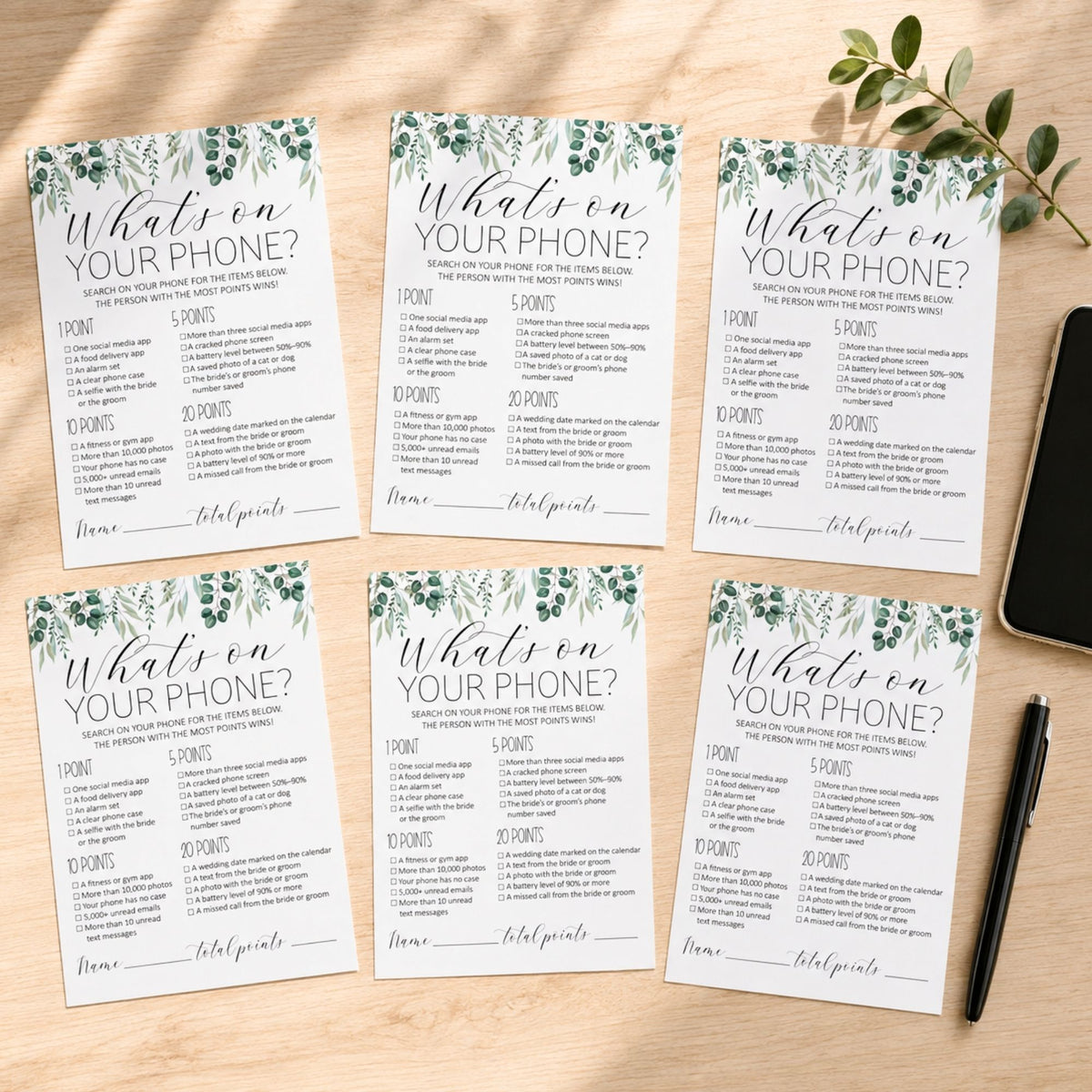 Six printable phone-themed party game cards arranged on a light wood surface, with a small green sprig and a black pen nearby. The cards read "What on YOUR PHONE?" and include scoring categories and fill-in lines.