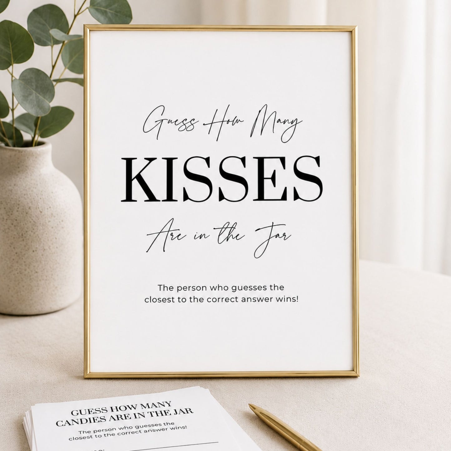Framed print with 'Guess How Many Kisses Are in the Jar' text on a light surface with a plant and paper in the background.