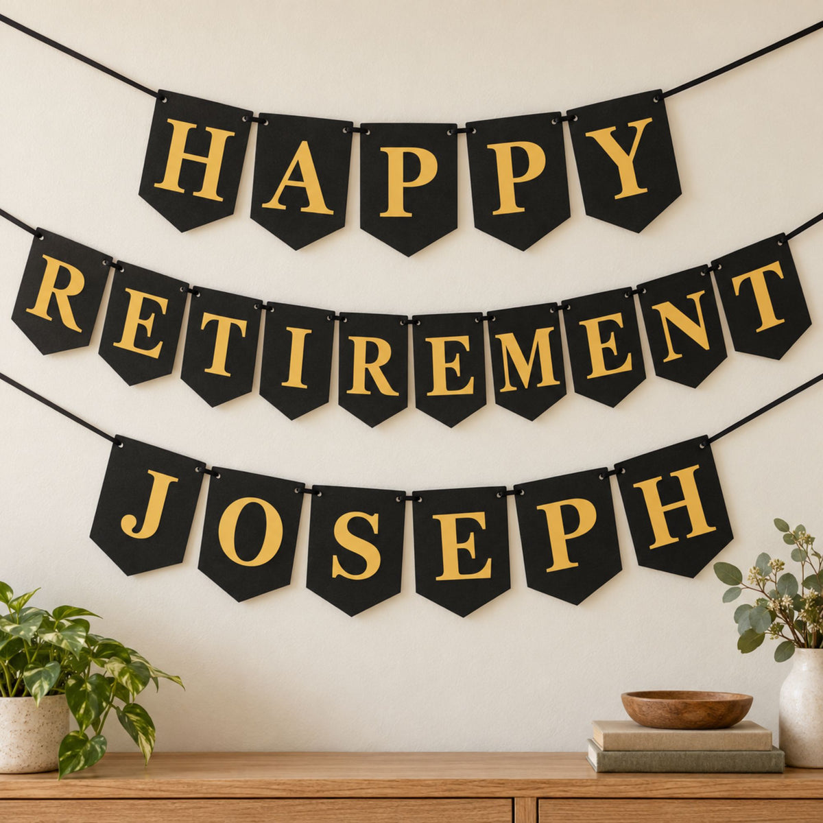 A black and gold personalized retirement banner with three rows of pennant reading 'HAPPY RETIREMENT JOSEPH' hanging on a wall above a wooden console table with plants and books.
