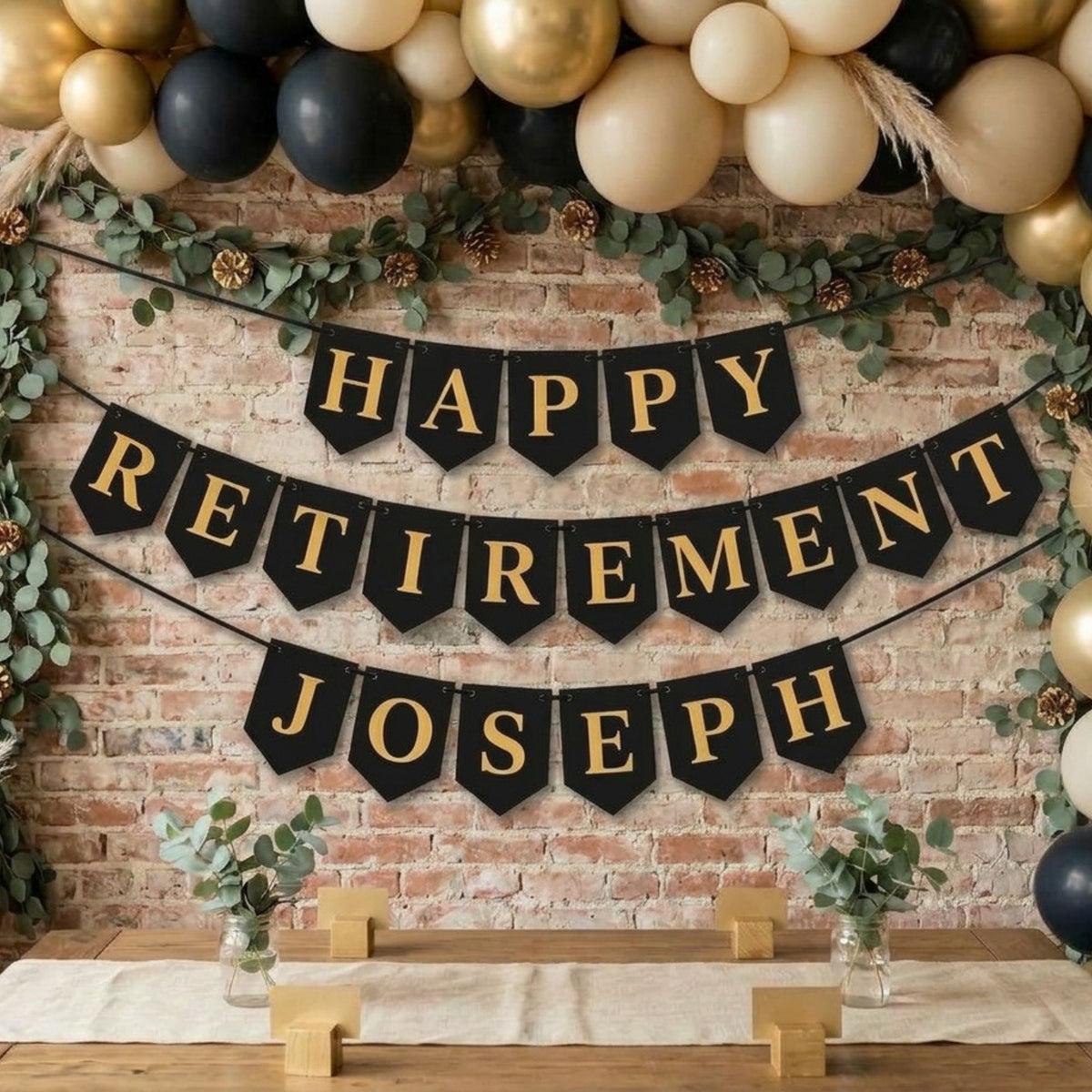 A festive retirement party backdrop featuring a black banner with gold lettering that reads 'HAPPY RETIREMENT JOSEPH' against a brick wall, decorated with black and gold balloons, green garlands, pinecones, and small plants on a table.
