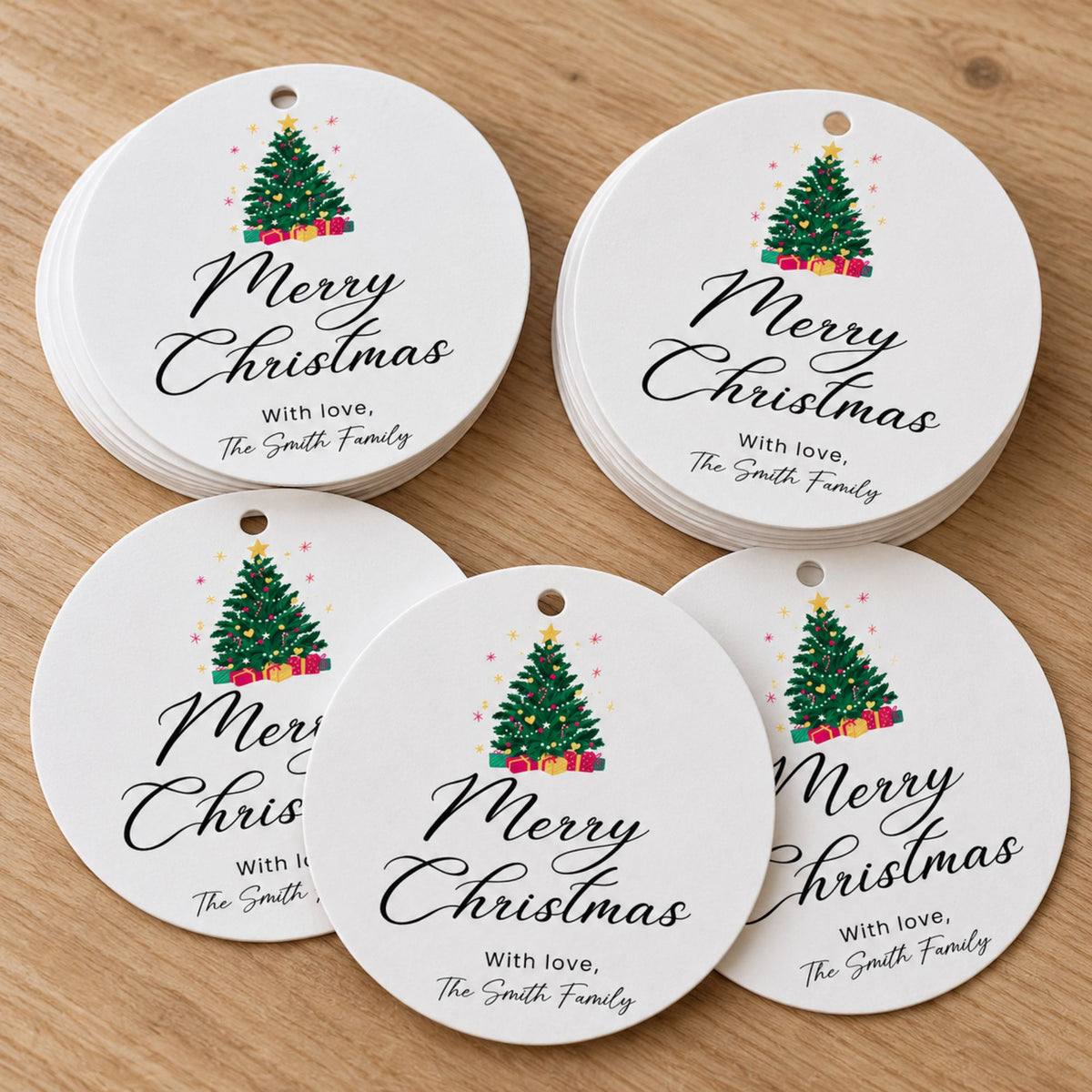 Five round white Christmas ornament tags with hanging holes, stacked on a light wood surface. Each tag features a colorful Christmas tree illustration and the printed message 'Merry Christmas With love, The Smith Family' in black script.