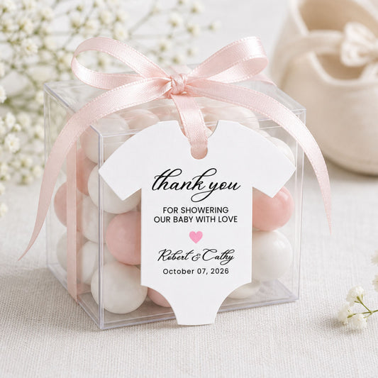 A clear square gift box filled with pastel baby shower candies in white and blush pink, tied with a pale pink satin ribbon and a baby onesie-shaped thank you tag. The tag reads 'thank you FOR SHOWERING OUR BABY WITH LOVE' with personalized names and a date.