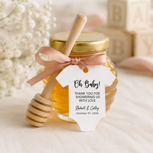 A glass honey jar with a gold lid, a wooden honey dipper resting on top, and a pale pink ribbon tied around the neck. A white baby onesie-shaped tag is attached with printed shower thank-you text.