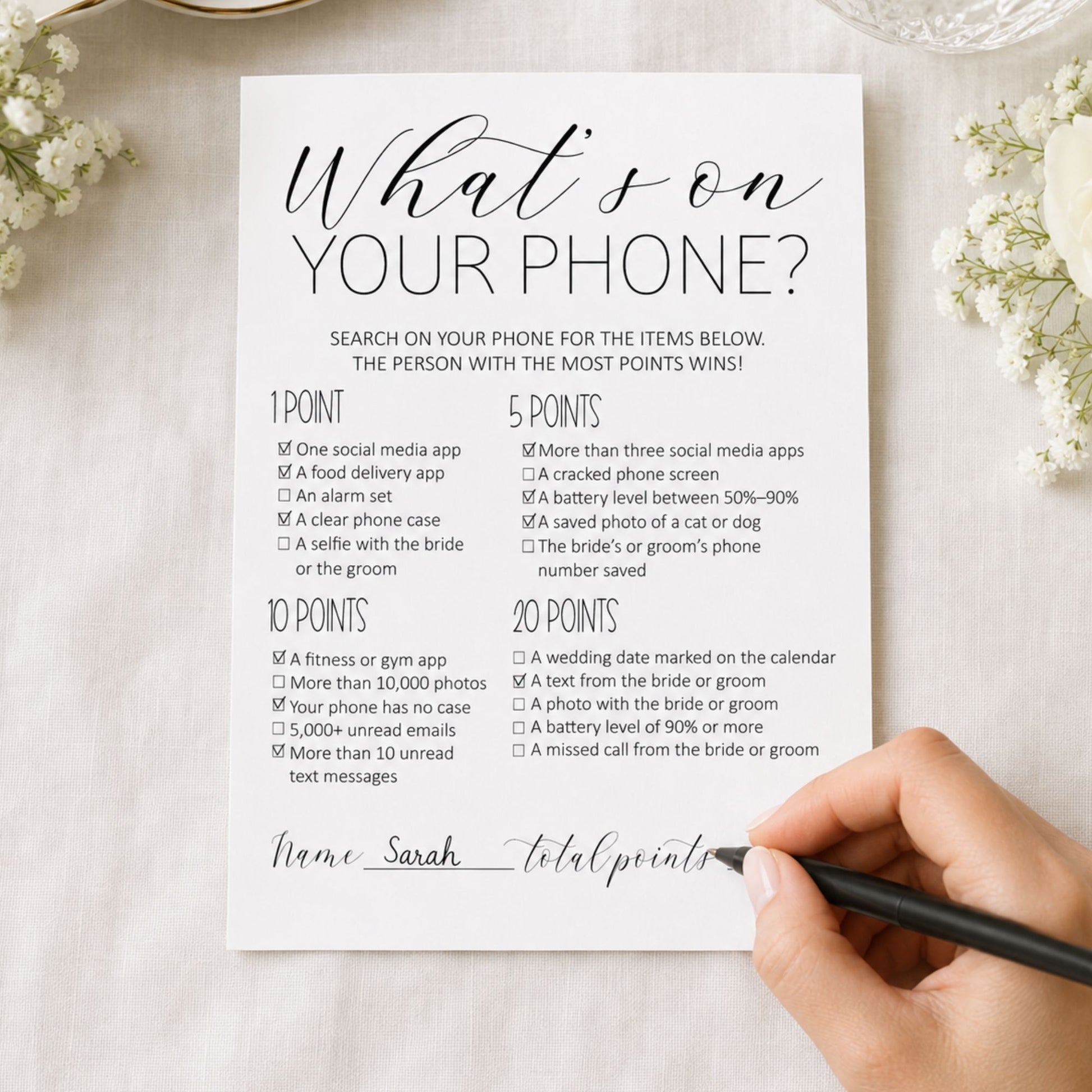 A printed novelty phone game card titled "What's on your phone?" shown on a light wood surface, with a hand holding a black pen near the bottom right. The card lists checkboxes and point values for phone-related items, with decorative flowers in the corners.